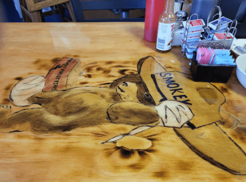 A wooden table featuring a detailed illustration of a bear wearing a hat, labeled "Smokey," holding a sign.